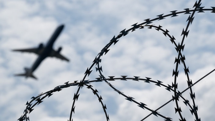 Ein Flugzeug am Himmel, im Vordergund Maschendrahtzaun.
