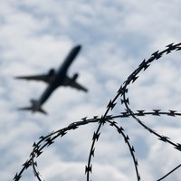 Ein Flugzeug am Himmel, im Vordergund Maschendrahtzaun.