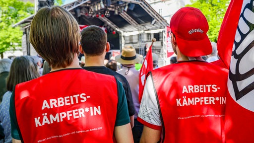 Zwei Besucher tragen Westen mit der Aufschrift "Arbeitskämpfer*in" auf dem Rücken