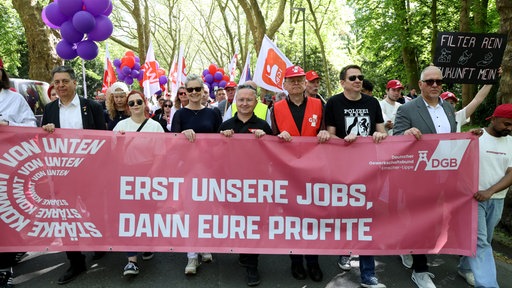 Am 1. Mai demonstrieren Menschen in Gelsenkirchen mit einem Plakat, auf dem „Erst unsere Jobs, dann eure Profite“ zu lesen ist