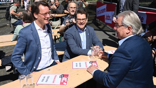 Hendrik Wüst (NRW Ministerpräsident), Thorben Albrecht (Vorsitzender des DGB Nordrhein-Westfalen) und Marc Buchholz (Oberbürgermeister in Mülheim an der Ruhr) sitzen auf einem Tisch auf der DGB Kundgebung zum 1. Mai