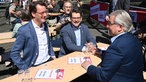 Hendrik Wüst (NRW Ministerpräsident), Thorben Albrecht (Vorsitzender des DGB Nordrhein-Westfalen) und Marc Buchholz (Oberbürgermeister in Mülheim an der Ruhr) sitzen auf einem Tisch auf der DGB Kundgebung zum 1. Mai