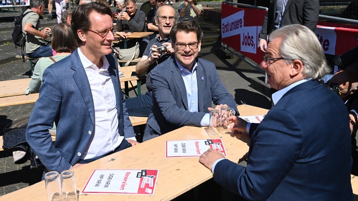 Hendrik Wüst (NRW Ministerpräsident), Thorben Albrecht (Vorsitzender des DGB Nordrhein-Westfalen) und Marc Buchholz (Oberbürgermeister in Mülheim an der Ruhr) sitzen auf einem Tisch auf der DGB Kundgebung zum 1. Mai