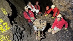 Sechs Höhlenforscher in Schutzanzügen mit einer Schubkarre voller Geröll