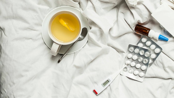 Eine Tasse Tee, Nasenspray, Medikamente und ein Fieberthermometer liegen auf einer Bettdecke
