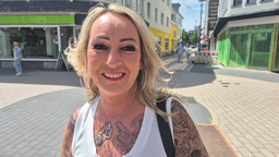 Eine Frau mit Tattoos und blonden Haaren steht auf einem Platz in einer Innenstadt.