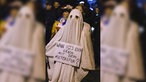 Eine als Geist verkleidete Person hält ein Schild mit der Aufschrift "Wann ist's euch genug mit dem Mietenspuk?"