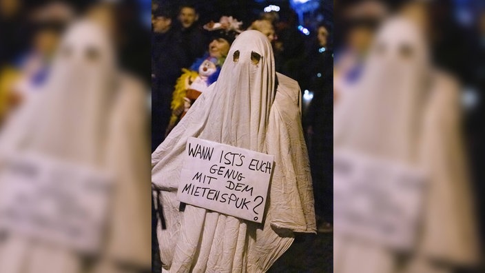 Eine als Geist verkleidete Person hält ein Schild mit der Aufschrift "Wann ist's euch genug mit dem Mietenspuk?"