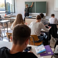 Symbolbild: Oberstufenschüler sitzen der Tafel zugewandt im Unterricht, die Lehrerin schreibt an die Tafel.