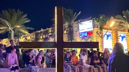 Ein Holzkreuz steht neben dem Megapark in einer nächtlichen Szene mit Feiernden im Hintergrund