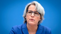 Porträtfoto einer Frau mit grauen kürzeren Haaren und Brille sitzt bei einer Pressekonferenz
