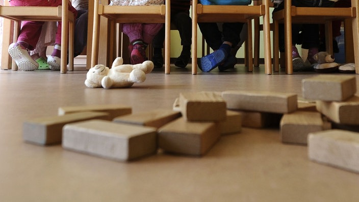 In einer Kita sind Bauklötze, ein Kuscheltier und Kinderbeine unter Stühlen zu sehen.
