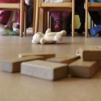 In einer Kita sind Bauklötze, ein Kuscheltier und Kinderbeine unter Stühlen zu sehen.