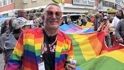 Winfried Kubitza-Simons mit Sakko in Regenbogenfagen beim CSD in Euskirchen neben einer großen Regenbogenflagge