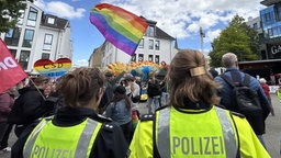 Zwei Polizistinnen stehen mit dem Rücken zur Kamera. Sie schauen auf eine große Regenbogenflagge
