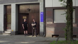 man sieht ein braunes Holztor, davor zwei Männer, die die Tür bewachen. 