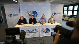 Vier Personen sitzen an einem Pult, hinter einem Polizeibanner auf einer Pressekonferenz