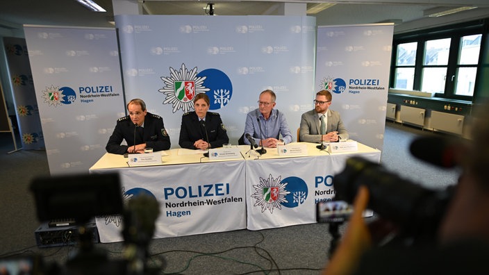 Vier Personen sitzen an einem Pult, hinter einem Polizeibanner auf einer Pressekonferenz