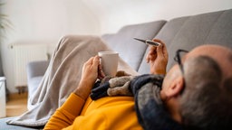 Eine Person liegt auf dem Sofa und schaut auf ein Fieberthermometer