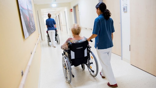 Zwei Pflegerinnen schieben in einem Klinikum Patienten mit Rollstühlen durch einen Flur.