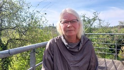 Frau mit weißen Haaren und Brille guckt in die Kamera, im Hintergrund eine grüne Landschaft