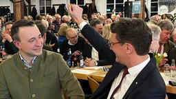 Hendrik Wüst sitzt an einer Tafel und lacht ins Publikum