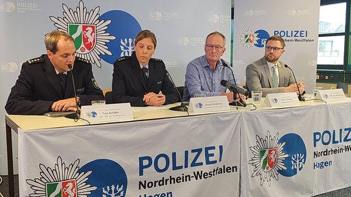 Vier Personen sitzen an einem Pult, hinter einem Polizeibanner auf einer Pressekonferenz