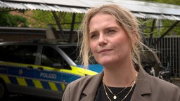 Eine blonde Frau bekleidet mit einem braunen Sakko steht vor einem Polizeiauto.