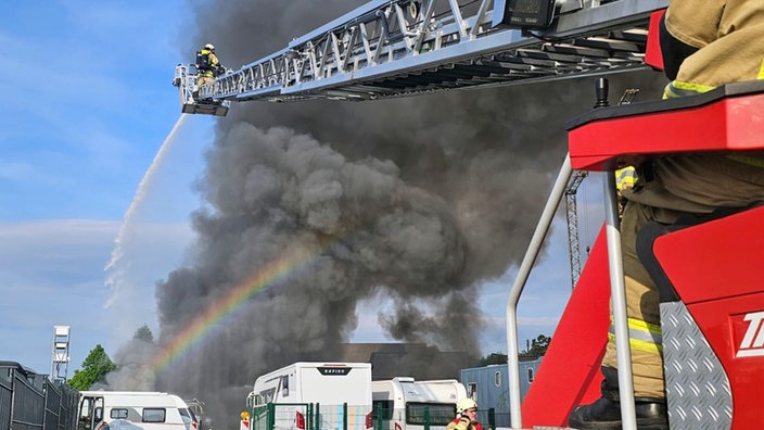 Eine große, schwarze Brandwolke über mehreren Wohnwägen. Die Einsatzkräfte der Feuerwehr stehen berits auf der Drehleiter und löschen den Brand.