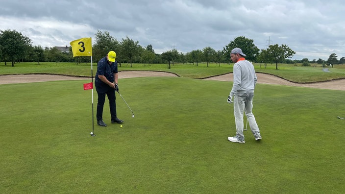 Zwei Männer schlagen auf einem Golfülatz den Ball in das Golfloch