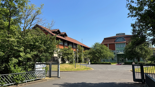 Eine Einfahrt führt auf einen Vorplatz vor zwei große Gebäude, die von Bäumen umgeben sind.