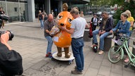 Die Maus wird auf einem Rollbrett von zwei Männern abtransportiert. Um sie herum stehen Menschen.