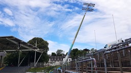 Ein schief stehender Flutlichtmast im Fußballstadion, links eine überdachte Tribüne mit grauen Stuhlreihen, rechts eine Stehtribüne