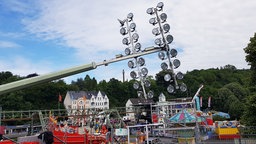 Schief liegender Flutlichmast über ein Kirmesgelände, im Hintergrund Häuser und das Schwebebahngerüst