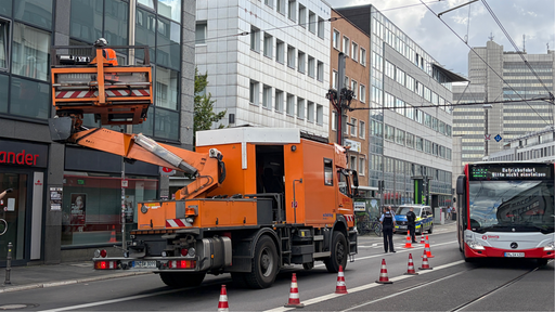 Ein orangenes Auto mit Krahn ist an einer Oberleitung über einer Hauptstraße zugange, ein Bus fährt drumherum.