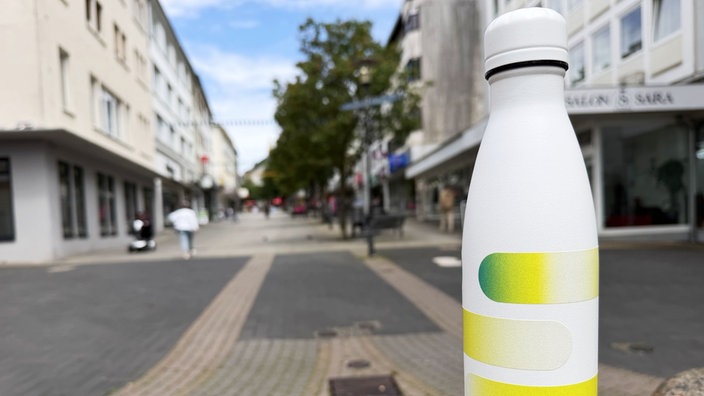 Eine weisse Trinkflasche mit gelbgrünen Streifen verziert, im Hintergrund unscharf eine Fußgängerzone
