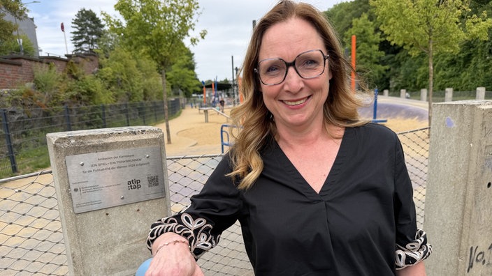 Eine Frau lehnt an einem Trinkwasserbrunnen, im Hintergrund ein Kinderspielplatz