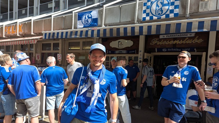 Manuel Steinhuber, Schalke Fan im blauen Schalke Trikot, steht bei Sonnenschein vor einer Kneipe und lacht.