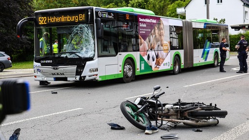 Bus mit Schaden an der Frontfensterscheibe steht vor einem stark demolierten Motorrad, umgeben von Polizisten