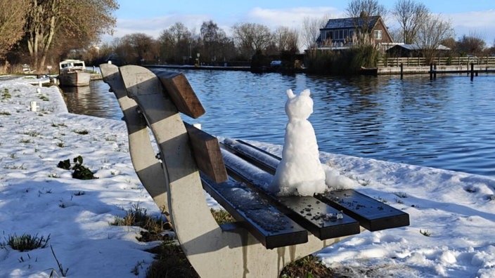 Eine aus Schnee gebaute Katze sitzt auf einer Parkbank und schaut auf ein Gewässer