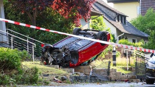 Ein rotes Auto liegt überschlagen in einem Vorgarten.