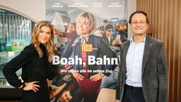 Ein blonde, schwarz gekleidete Frau neben einem großen Plakat, das sie in Bahnkleidung zeigt