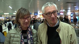 Ein älteres Paar, beide mit Brille und in beigen Jacken in der Bahnhofshalle
