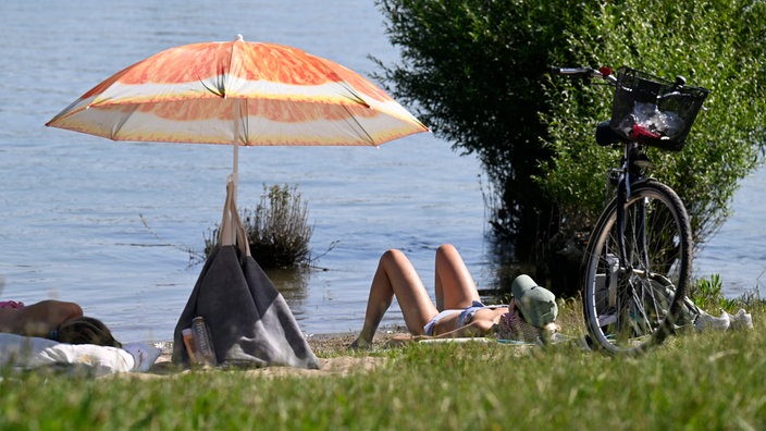Rodenkirchen in NRW: Eine Frau liegt am Ufer des Rheins neben einem Sonnenschirm.