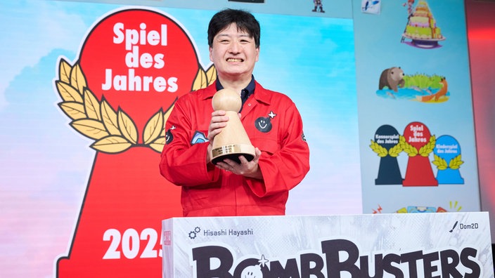 Der japanische Autor Hisashi Hayashi hält im roten Overall gekleidet die Auszeichnung für das "Spiel des Jahres in die Höhe".