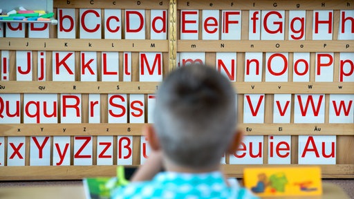 Ein Kind sitzt an einem Tisch; im Hintergrund sind Buchstaben zu sehen