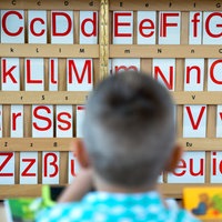 Ein Kind sitzt an einem Tisch; im Hintergrund sind Buchstaben zu sehen