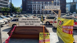 Auf dem „Parkplatz Friedensplatz“ vor dem Dortmunder Rathaus stehen viele Taxis. Im Vordergrund ist ein Sarg auf einem Anhänger zu sehen. Daruf steht ein Schild mit der Aufschrift: "Ohne Mndestpreis beerdigen die Behörden DAS TAXI"