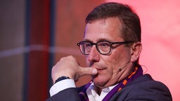 Ein Mann mit Brille hält sich den Daumen an den Mund und guckt nachdenklich