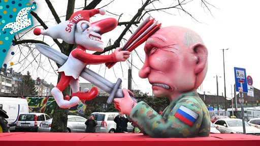 Putin ersticht eine Figur auf der "Satire" steht. Diese haut ihm mit einem Fächer auf den Kopf